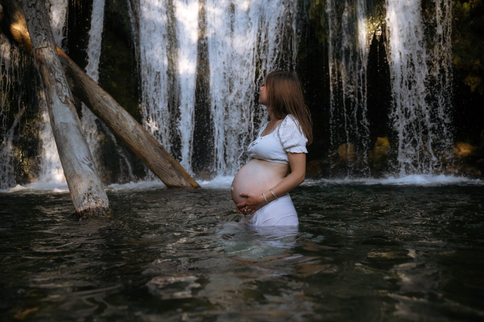 Femme enceinte de profil dans l'eau devant une cascade, robe blanche, mains sur le ventre