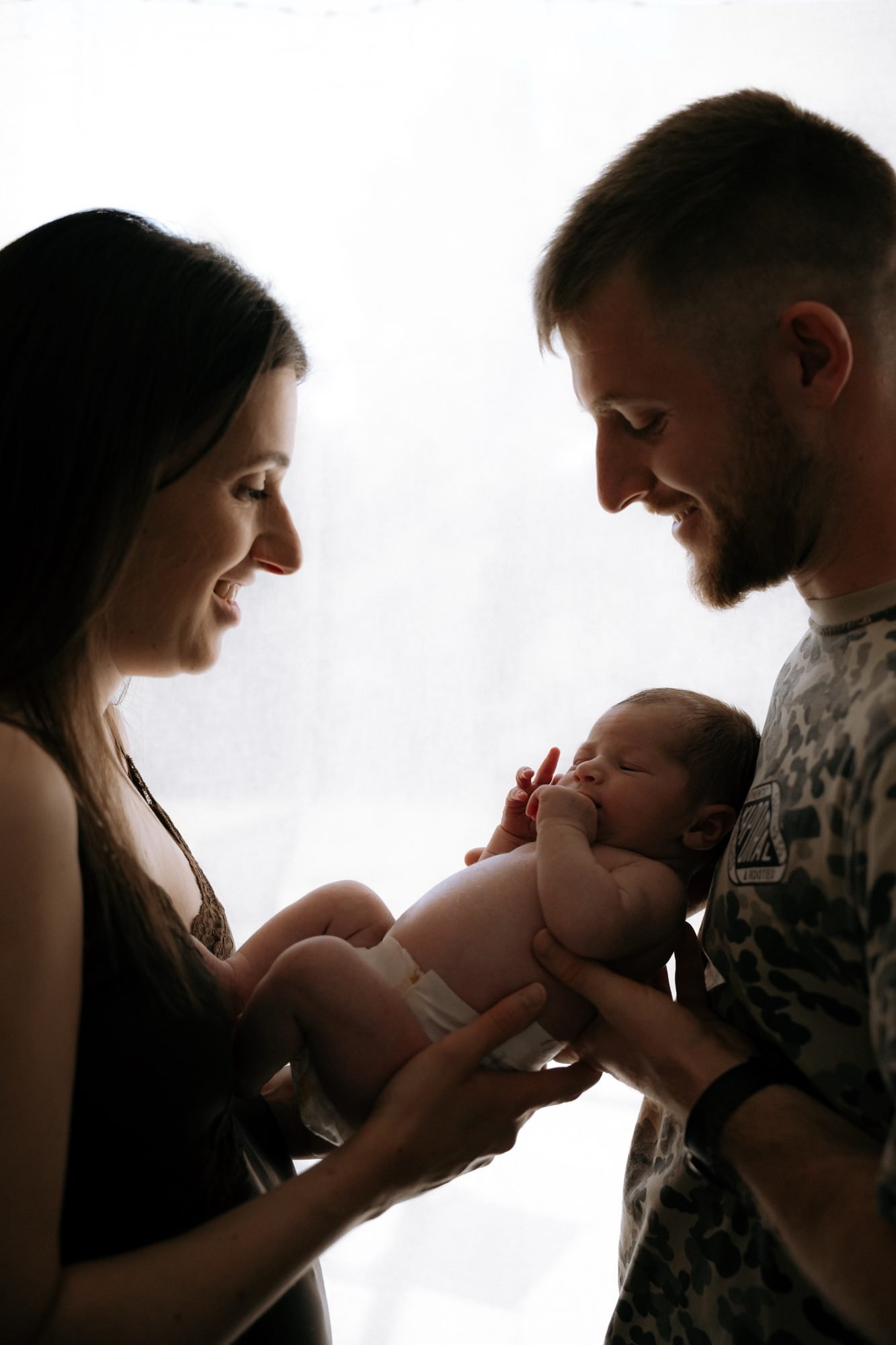 Deux parents tenant leur nouveau-né, en contre-jour près d'une fenêtre